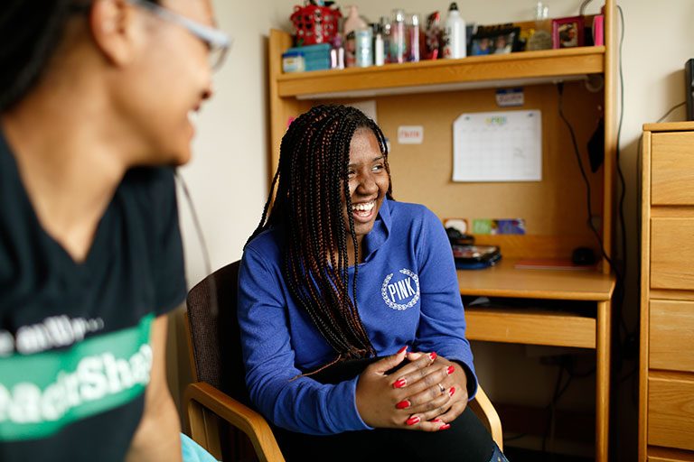 Two young women laugh in a dorm room.