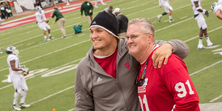 A young man and an older man stand on the IU football field smiling with players in the background.