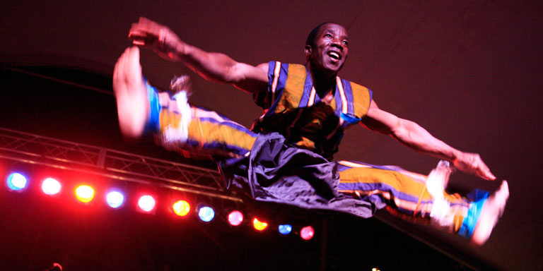 A man in colorful costume does a split toe-touch jump.