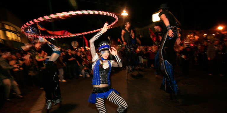 A woman in a costume twirls a hula hoop above her head as other costumed performers on stilts walk nearby.