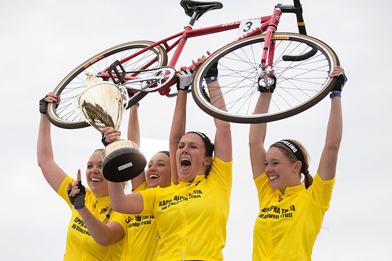 A winning women's Little 500 team holds a bike and trophy above their heads.